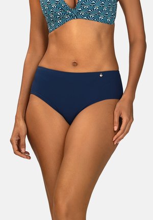 Kobieta ubrana w niebieski bikini z kwiatowym wzorem na górze i jednolite granatowe bikini z wysokim stanem na dole, stojąca na tle jednolitej białej ściany.