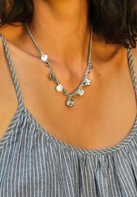 Collana d'argento con vari ciondoli, tra cui forme rotonde e floreali, appesi a una sottile catena, indossata sopra un top a righe grigie.