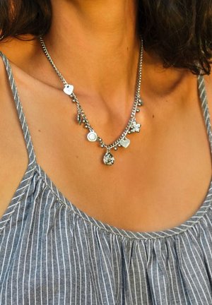 Silberne Halskette mit verschiedenen Charms, darunter runde und florale Formen, die an einer dünnen Kette hängen, getragen über einem grau gestreiften Oberteil.