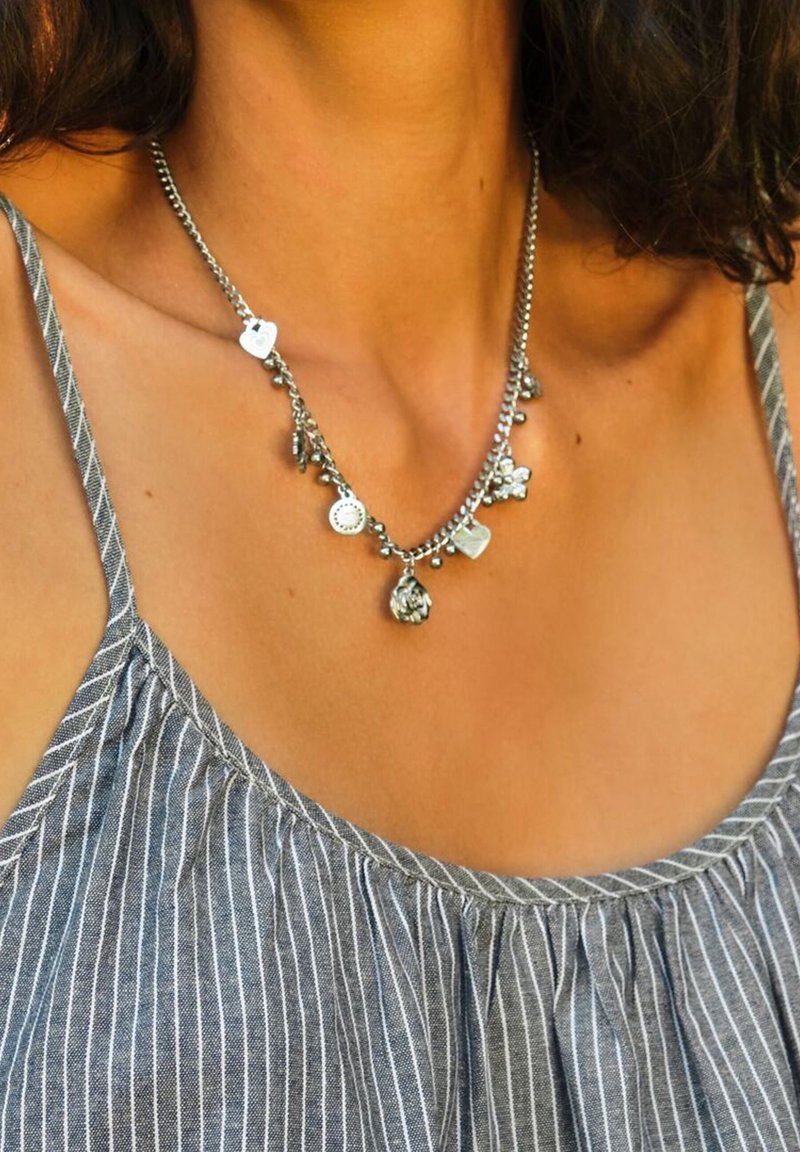 Collana d'argento con vari ciondoli, tra cui forme rotonde e floreali, appesi a una sottile catena, indossata sopra un top a righe grigie.