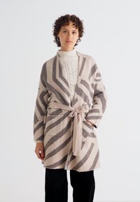 Beige mintás kardigán barna zigzag csíkokkal, hosszú ujjal, deréknál megkötő övvel és két elülső zsebbel. Tejfehér kötött pulóver fölött viselve.