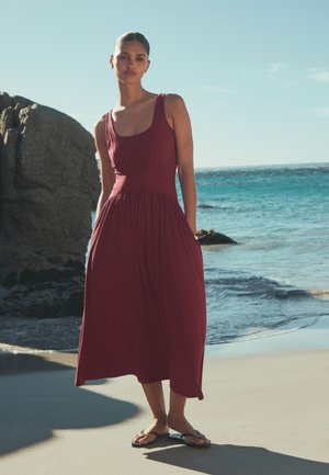 Sieviete bez piedurknēm sarkanbrūnā kleitā un sandales uz kājām stāv uz smilšainas pludmales pie okeāna un liela akmens skaidrā debesīs.