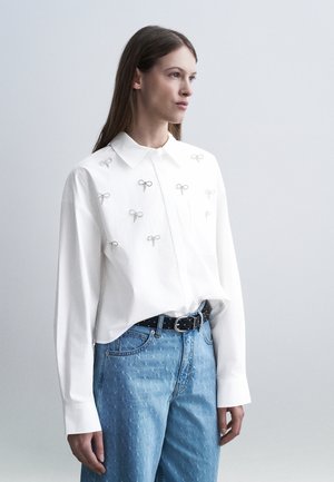 Kvinne iført en hvit langermet skjorte med små broderte sløyfer, kombinert med høytlivs blå jeans og et svart belte.