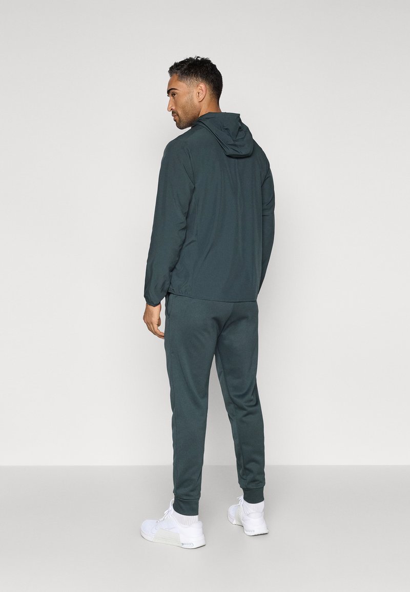 Chándal con capucha en verde oscuro, fabricado con un tejido ligero. Presenta puños y dobladillo elásticos, combinado con zapatillas deportivas blancas.