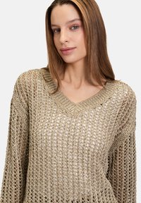 Pull en tricot de couleur or clair avec un tissage lâche et ouvert. Caractéristiques d'un décolleté en V et de manches larges. La texture est douce et légèrement transparente.