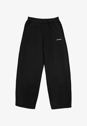 Persoon draagt zwarte broek met wijde pijpen met witte geborduurde tekst en zwarte schoenen, houdt een donker jasje in één hand.