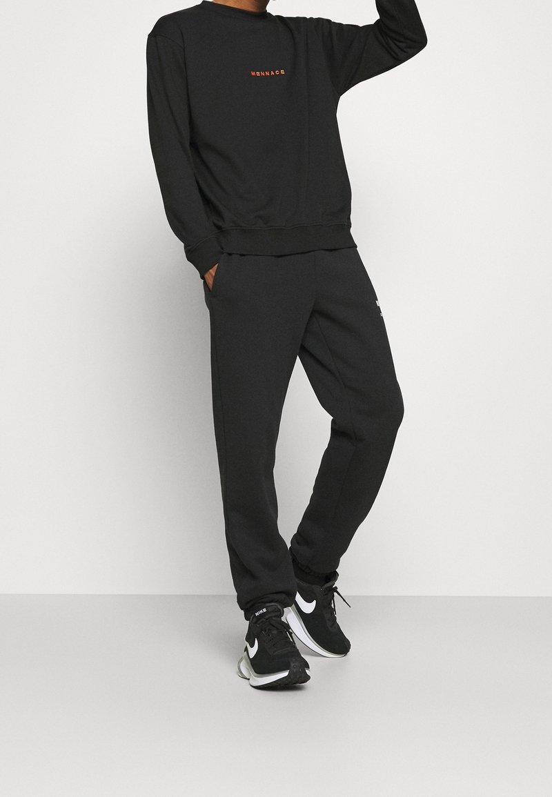 Personne portant un sweat-shirt noir avec le texte orange "MENNACE", un pantalon de jogging noir et des baskets Nike noires avec des logos swoosh blancs.