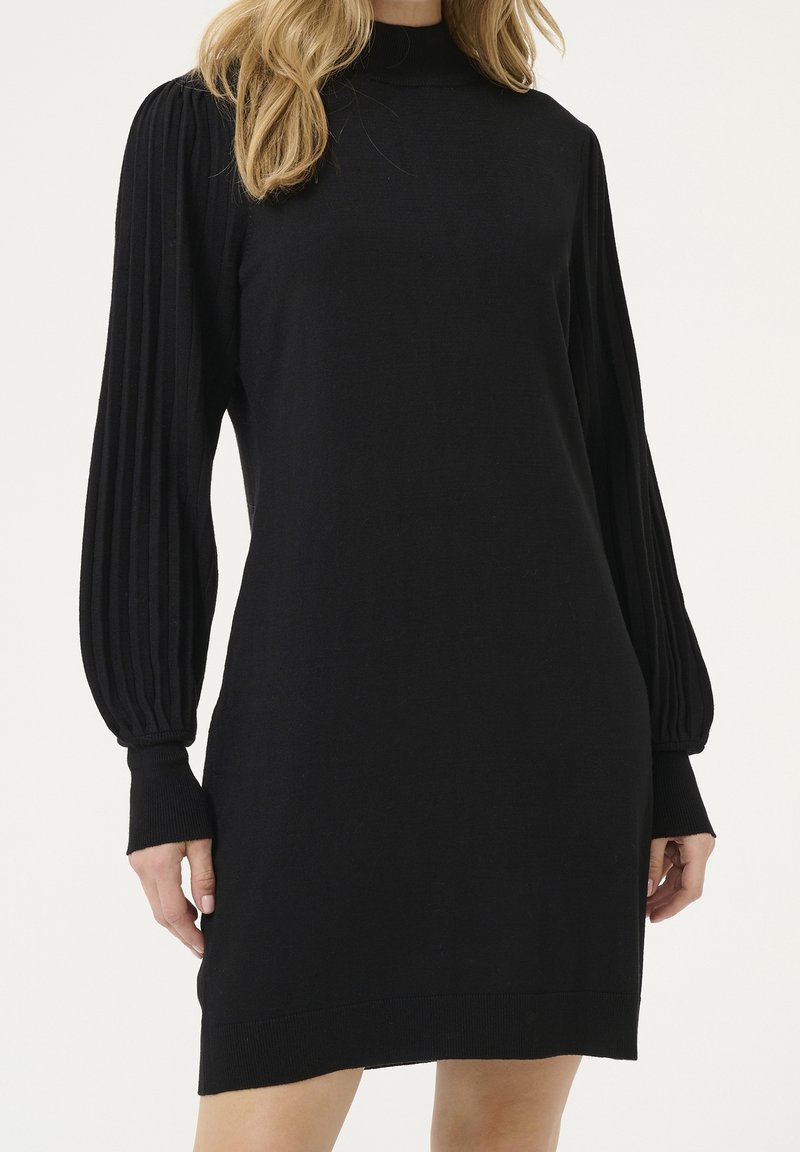 Femme portant une robe en maille noire avec des manches longues plissées et des poignets côtelés, debout devant un fond clair uni.