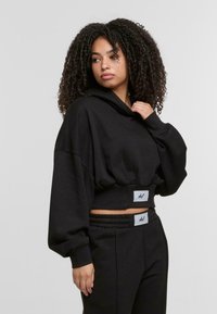Zwarte cropped hoodie van zacht materiaal met een capuchon en brede mouwen, met een opvallend logo op de tailleband, gecombineerd met bijpassende zwarte broeken.