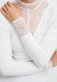 Hvid langærmet top med høj blondekrave og blonde-accenterede manchetter, med en ribbet struktur og indviklede blonde mønstre.