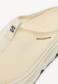Ljusbeige Salomon slip-on sko med ovandel i mesh, sydda detaljer, logotyp och en texturerad sula i kontrasterande svart.