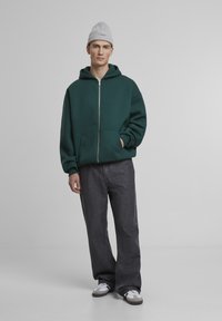 Grøn zip-up hoodie med store forlommer og en afslappet pasform, parret med mørkegrå vide bukser og hvide sko, sat mod en ensfarvet baggrund.