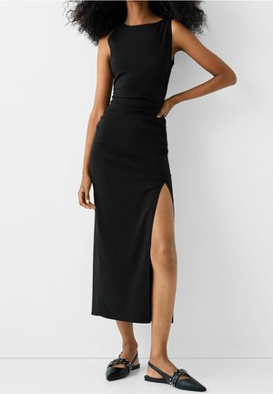 Femme portant une robe noire sans manches avec une fente haute sur le côté et des chaussures plates noires cloutées, debout sur un fond blanc uni.