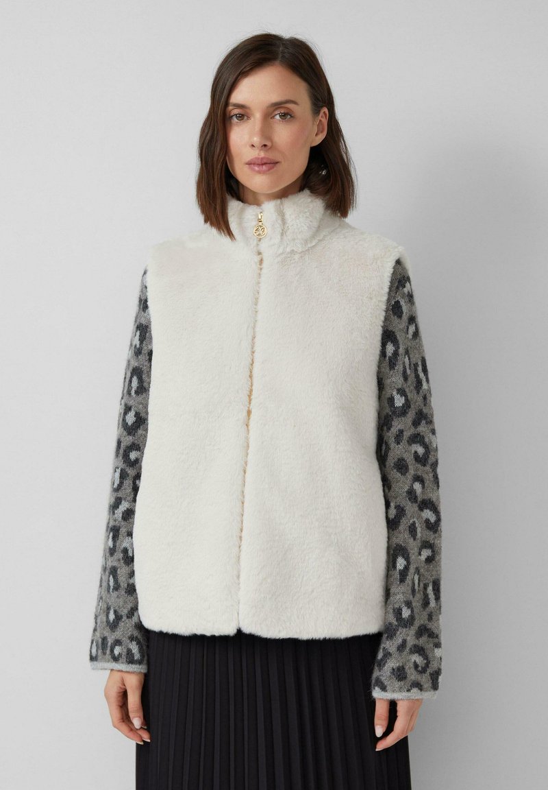 Eine Frau trägt eine weiße, flauschige Weste über einem grauen Leopardenmuster-Pullover und einem schwarzen Faltenrock, während sie vor einem schlichten, hellen Hintergrund steht.