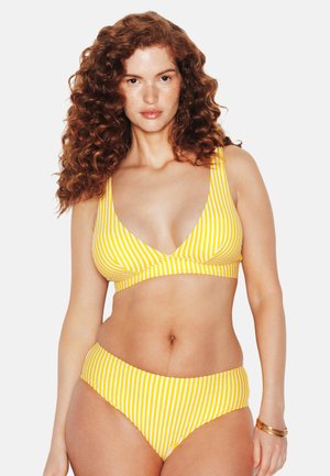 Femme aux cheveux roux bouclés portant un bikini rayé jaune et blanc, debout devant un fond blanc avec une expression neutre.