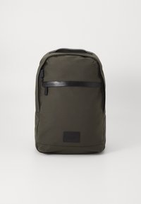 Zaino verde scuro realizzato in tessuto testurizzato, con un accento in pelle nera sulla cerniera e un'etichetta rettangolare con logo nella parte inferiore.