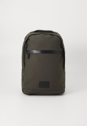 Zaino verde scuro realizzato in tessuto testurizzato, con un accento in pelle nera sulla cerniera e un'etichetta rettangolare con logo nella parte inferiore.