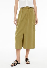 Khaki mid-length krilo z elastičnim pasom, dvema stranskima žepoma, sprednjim razporkom in gladko teksturo. V kombinaciji s črnimi trakastimi sandali.
