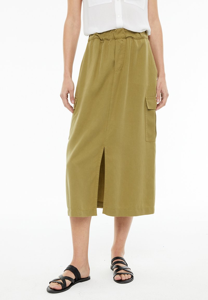 Khaki mid-length krilo z elastičnim pasom, dvema stranskima žepoma, sprednjim razporkom in gladko teksturo. V kombinaciji s črnimi trakastimi sandali.