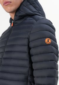 Navy puffer jacket with horizontal quilting, a zipper front, and an orange accent at the collar and a logo badge on the sleeve.