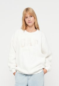 Felpa in pile bianco con vestibilità ampia, caratterizzata dal logo "GAP" in rilievo, polsini a coste e scollo rotondo. Abbinata a jeans in denim chiaro.