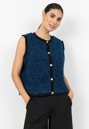 Kobieta o ciemnych włosach związanych w kucyk nosi niebieską, teksturowaną kamizelkę bez rękawów z złotymi guzikami i czarnym obszyciem, w parze z czarnymi spodniami, z rękami w kieszeniach.