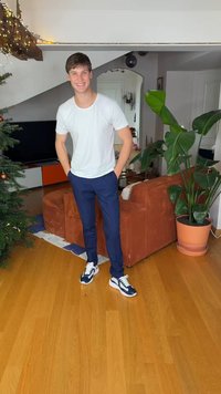 Hvid T-shirt, marineblå bukser og marineblå sneakers med hvide snørebånd. Indendørs omgivelser med brun sofa og potteplante i baggrunden.