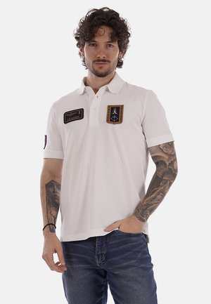 Uomo con capelli ricci che indossa una polo bianca con toppe e jeans blu, in piedi con una mano in tasca contro uno sfondo semplice.