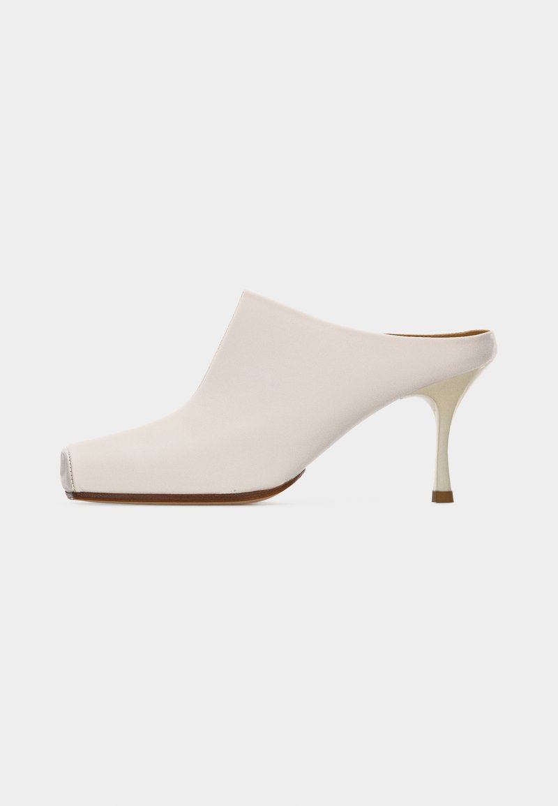Μπεζ μουλ με μυτερή μύτη και λεπτό τακούνι στιλέτο μέτριου ύψους, εμφανιζόμενο από το πλάι σε απλό φωτεινό φόντο.