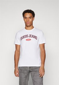 Baltos trumpomis rankovėmis medvilninės marškinėliai su raudonu "GUESS JEANS 1981" grafiniu spaudiniu priekyje. Derinama su pilkais džinsiniais kelnėmis.