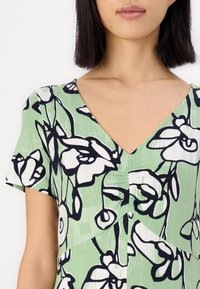 Rochie cu decolteu în V, dintr-un material verde deschis cu un model floral alb și negru, având mâneci scurte și detalii plisate la bust.