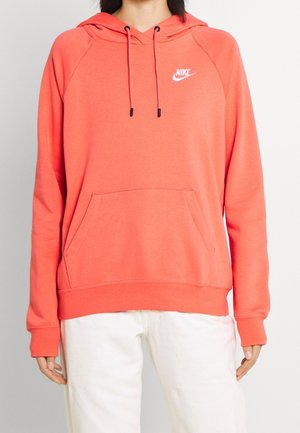 Personne portant un sweat à capuche Nike corail avec poche kangourou et cordon de serrage, associé à un pantalon blanc sur un fond uni.