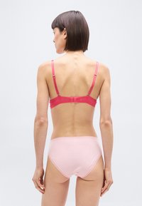 Γυναίκα με κοντά σκούρα μαλλιά, σε θέα από πίσω, φορώντας ροζ σουτιέν και ανοιχτό ροζ εσώρουχο με ψηλή μέση, μπροστά σε απλό λευκό φόντο.