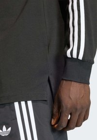 Čierna dlhá košeľa s bielymi bočnými rozparkami a tromi bielymi pruhmi na rukáve. Bavlnený materiál, priliehavý strih. Ruka je viditeľná.