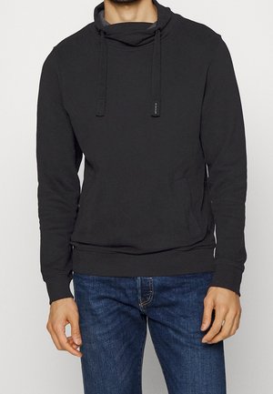 Mann trägt schwarz Kapuzenpullover mit aufgesetzter Tasche vorne und blaue Jeans, steht vor einfachem hellem Hintergrund.