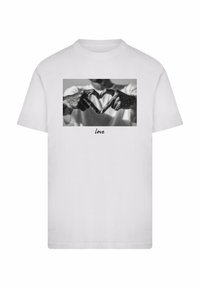 Hvid bomulds T-shirt med et gråtonet grafisk motiv af hænder, der danner en hjerteform, med ordet "kærlighed" trykt under billedet.