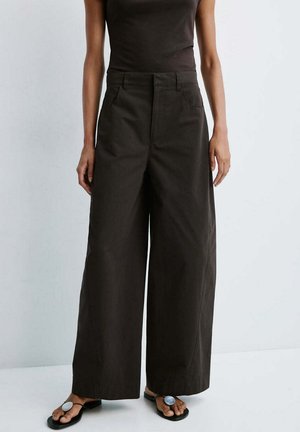 Mujer con pantalones negros de cintura alta y pernera ancha y sandalias negras con grandes adornos circulares plateados en los dedos.