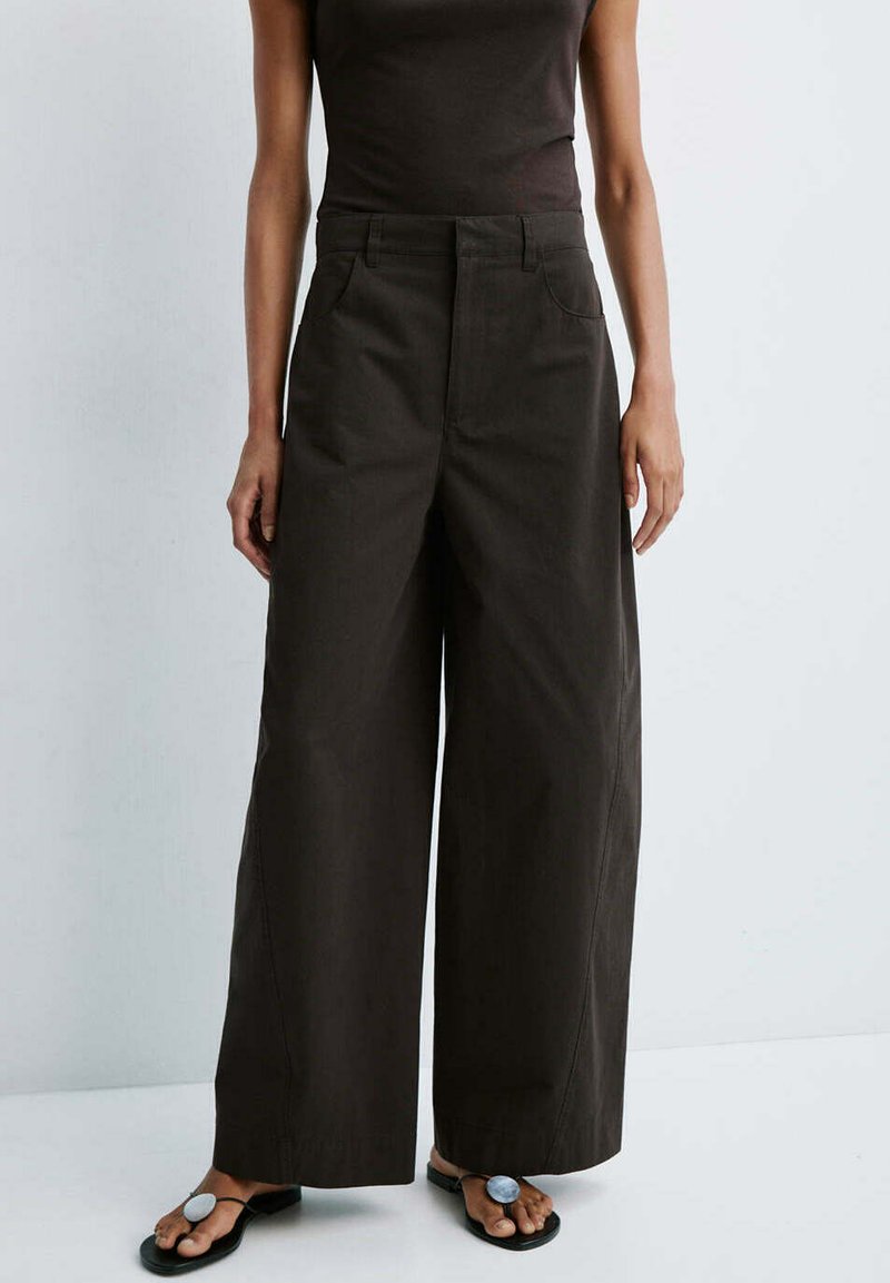 Femme portant un pantalon noir taille haute à jambes larges et des sandales noires avec de grandes embellissements argentés circulaires sur les orteils.
