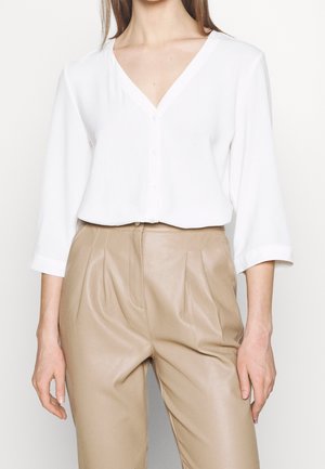 Woman wearing a white V-neck buttoned blouse with 3/4 sleeves tucked into high-waisted beige pleated leather pants.