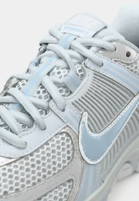 Close-up de um sneaker Nike Vomero 5 azul e branco, mostrando a textura da malha, os atacadores e o logo swoosh na lateral.