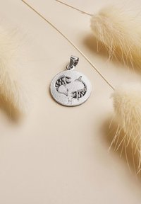 Rund silverhänge med graverade två stiliserade fiskar som vänder sig mot varandra, placerad på en beige yta med mjuka, beige torra blommor i närheten.