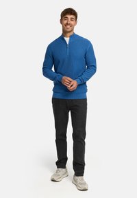 Blauer Zip-Kragen-Pullover aus strukturiertem Strickmaterial, kombiniert mit dunklen Jeans und beigen Sneakern. Der Pullover hat gerippte Bündchen und einen gerippten Saum.