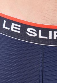 Caleçons boxers bleu marine avec un tissu texturé, dotés d'une taille noire avec des accents rouges et blancs et le texte "LE SLIP" affiché en évidence.