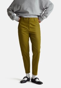 Pantaloni verdi con una texture liscia, forma affusolata e chiusura con bottone in vita, abbinati a una felpa grigia e scarpe nere.