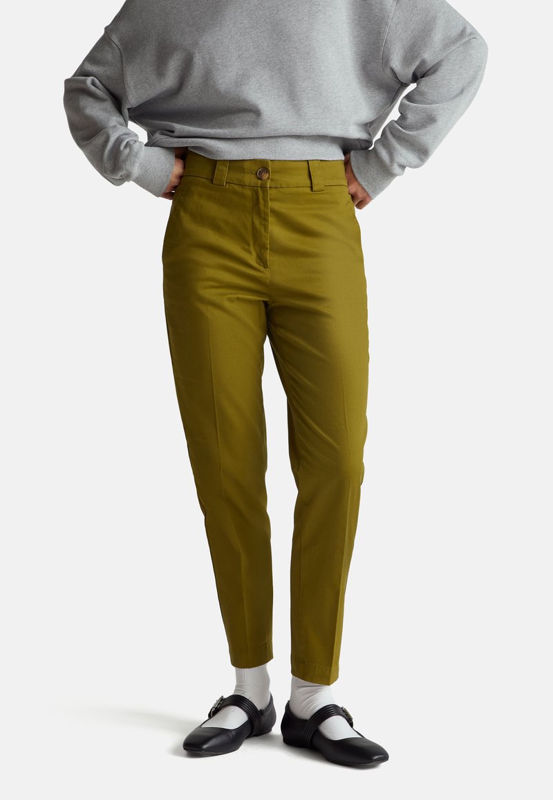 Pantaloni verdi con una texture liscia, forma affusolata e chiusura con bottone in vita, abbinati a una felpa grigia e scarpe nere.