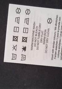 Hoito-ohjemerkki, jossa mustat kuvakkeet valkoisella kangaspohjalla; sisältää ohjeet pesuun, kuivaamiseen ja silittämiseen.