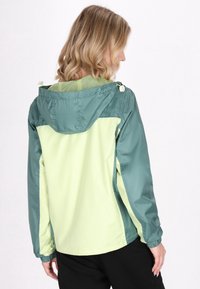 Chaqueta cortavientos verde y amarillo claro con capucha, presenta puños elásticos y un interior de malla, vista desde la parte trasera.