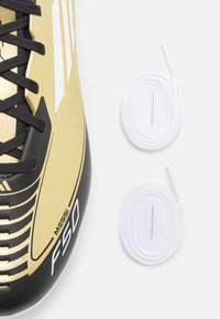 La scarpa da calcio oro e nera presenta un design elegante con superficie texturizzata, tre strisce bianche e include due lacci bianchi.
