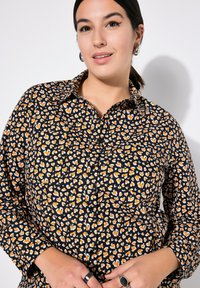Vrouw met donker haar in een staart, gekleed in een zwart, oranje en wit overhemd met hartpatroon, en zilveren creolen en ringen.