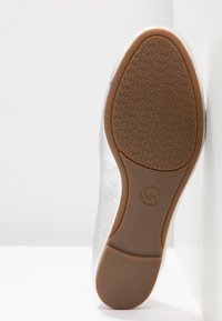 Semelle d'une chaussure plate argentée avec une semelle en caoutchouc marron clair présentant des motifs répétés du logo "MK" et un logo circulaire "Michael Kors".